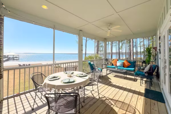A screened-in porch with a wooden floor features an outdoor dining table set for four and a separate seating area with a teal sofa, all overlooking a scenic waterfront view.