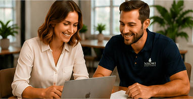 Two professionals collaborate at a wooden table, smiling while looking at a laptop. Plants adorn the bright, modern workspace, suggesting a productive atmosphere.