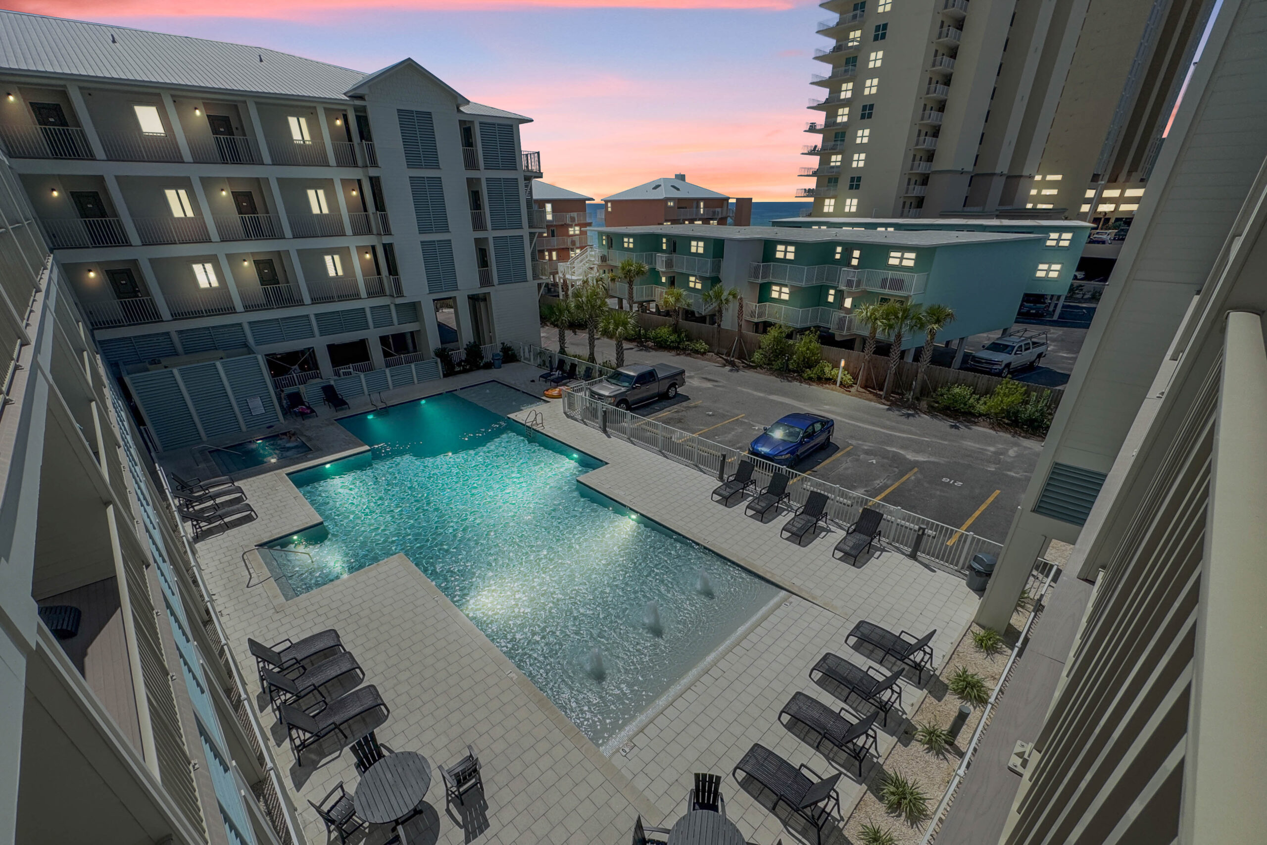 Aerial view of a hotel pool surrounded by lounge chairs, with multiple buildings and a sunset in the background, inviting relaxation.