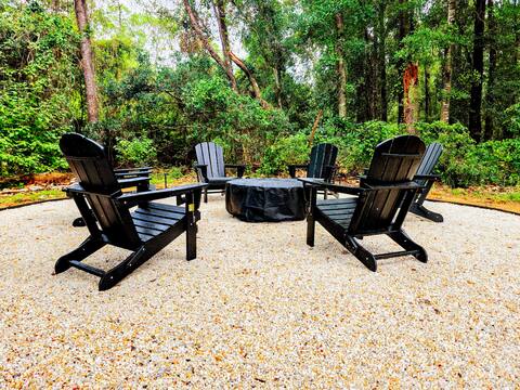 Five black Adirondack chairs arranged in a circular pattern around a covered fire pit, set on a gravel area amidst lush green trees.