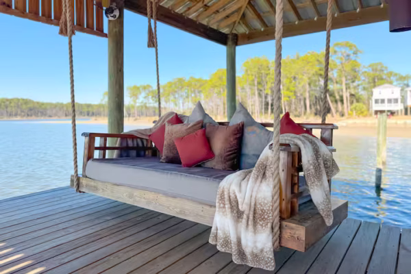 A large covered boathouse with a metal roof sits over the water, featuring a lounge area with outdoor seating and an attached boat slip housing a motorized boat.A spacious covered wooden boat dock offers comfortable seating, a swinging daybed, and panoramic views of a wide, calm body of water.A large wooden daybed swing, layered with decorative pillows and plush blankets, hangs from thick ropes under a covered boat dock overlooking calm water.