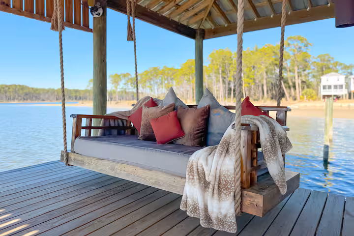 A large covered boathouse with a metal roof sits over the water, featuring a lounge area with outdoor seating and an attached boat slip housing a motorized boat.A spacious covered wooden boat dock offers comfortable seating, a swinging daybed, and panoramic views of a wide, calm body of water.A large wooden daybed swing, layered with decorative pillows and plush blankets, hangs from thick ropes under a covered boat dock overlooking calm water.