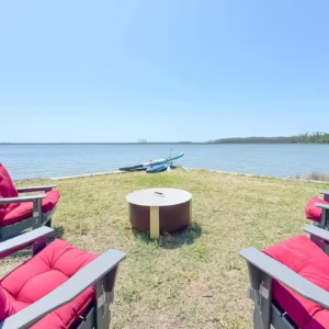Four red cushioned Adirondack chairs surround a central fire pit on a grassy lawn overlooking a wide body of water.