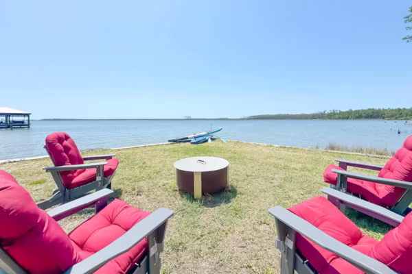 Four red cushioned Adirondack chairs surround a central fire pit on a grassy lawn overlooking a wide body of water.