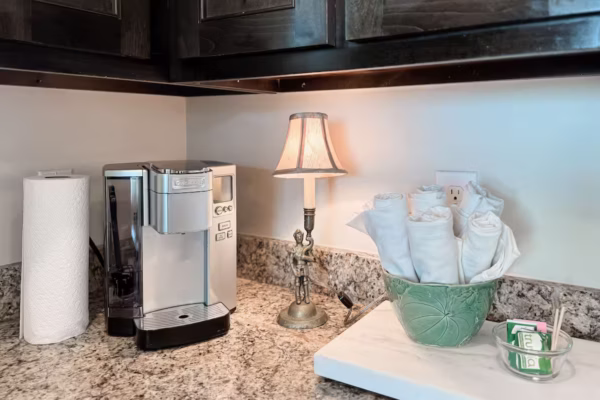 A kitchen coffee station features a single-serve coffee maker, a roll of paper towels, a small decorative lamp, and a green bowl filled with rolled white linens on a granite countertop.