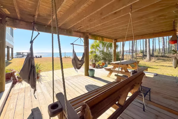 A covered wooden deck features a picnic table, an outdoor charcoal grill, and views of a sunny lawn, pine trees, and a calm waterfront.