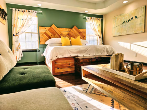 A cozy bedroom with sage green walls featuring a wooden headboard and matching platform bed with white bedding and two yellow pillows. In the foreground is a dark green upholstered bench and a wooden coffee table resting on a patterned rug, with sunlight streaming through windows on either side of the bed.