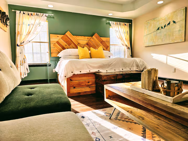 A cozy bedroom with sage green walls featuring a wooden headboard and matching platform bed with white bedding and two yellow pillows. In the foreground is a dark green upholstered bench and a wooden coffee table resting on a patterned rug, with sunlight streaming through windows on either side of the bed.