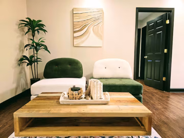 A stylish living room features two low-profile armchairs—one with a white base and a dark green backrest, and the other with a dark green base and a white backrest—positioned in front of a neutral wall with an abstract beige and white painting. A large wooden coffee table sits in the foreground with a tray holding decor, while a tall potted plant stands to the left and a black door is visible on the right.