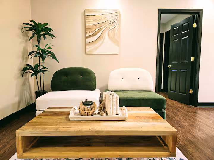 A stylish living room features two low-profile armchairs—one with a white base and a dark green backrest, and the other with a dark green base and a white backrest—positioned in front of a neutral wall with an abstract beige and white painting. A large wooden coffee table sits in the foreground with a tray holding decor, while a tall potted plant stands to the left and a black door is visible on the right.
