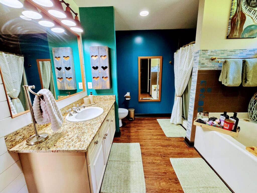 Brightly colored bathroom featuring a double sink vanity with granite countertop, decorative heart-shaped wood art, and a relaxing bathtub setup.