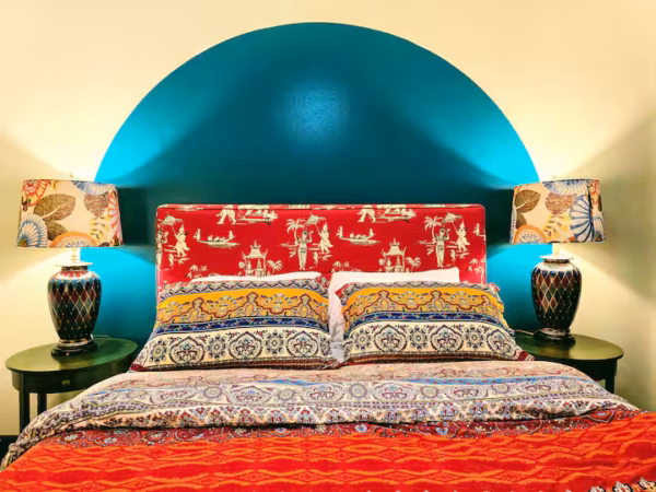 A vibrant bedroom featuring a bed with a red patterned headboard and colorful patterned pillows, set against a wall with a bold teal semi-circular painted backdrop. Matching nightstands on each side hold patterned lamps with matching lamp shades.