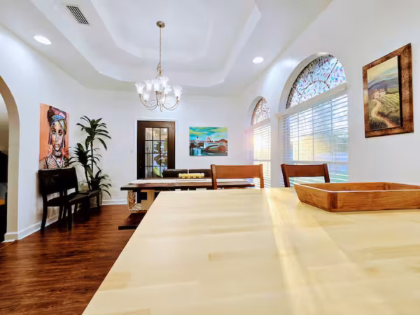 A view from a light wood kitchen island countertop looking across into an open-concept dining area featuring a dark wooden dining table, a traditional chandelier suspended from a tray ceiling, arched windows, and colorful artwork on the white walls.