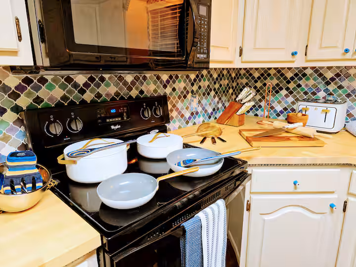 A close-up kitchen view featuring a black electric range and microwave, cream-colored raised-panel cabinets with bright blue knobs, a colorful lantern-pattern mosaic tile backsplash, and light wood countertops arranged with white cookware and a knife block.