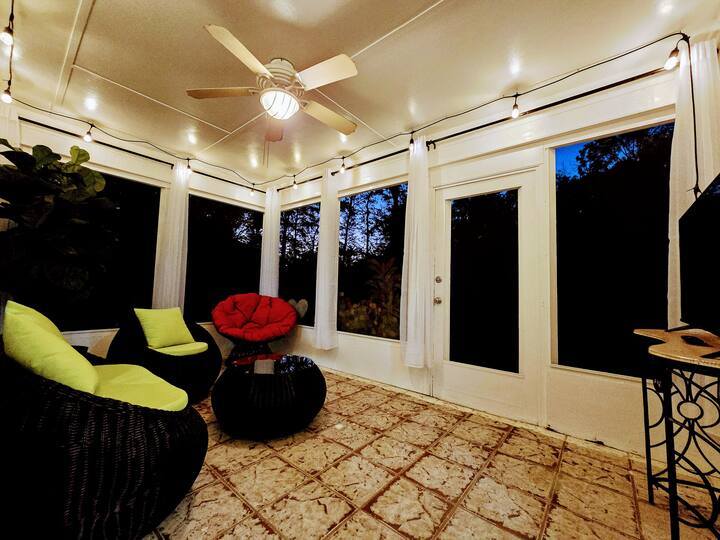 Cozy sunroom with modern furnishings, vibrant green and red cushions, string lights, and large windows overlooking a wooded area.