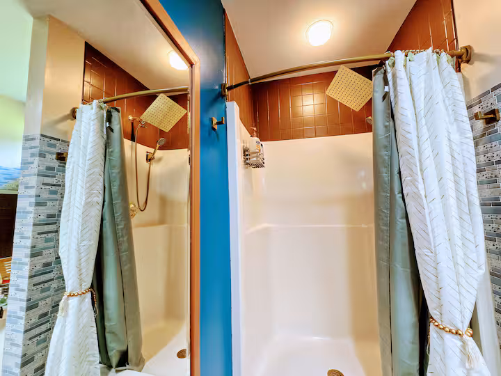 A brightly lit bathroom features two side-by-side shower stalls equipped with white-and-green patterned curtains, brass fixtures, and square overhead rain shower heads.