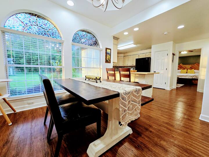 Bright dining area featuring a wooden table with a decorative runner, surrounded by chairs. Large arched windows allow natural light, revealing a garden view. The open kitchen is visible in the background.