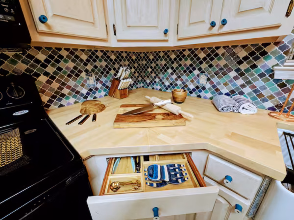 A close-up view of a kitchen corner featuring light wood butcher block countertops, cream-colored cabinets with blue knobs, an open drawer showcasing organized kitchen utensils and oven mitts, and a colorful lantern-pattern mosaic tile backsplash.