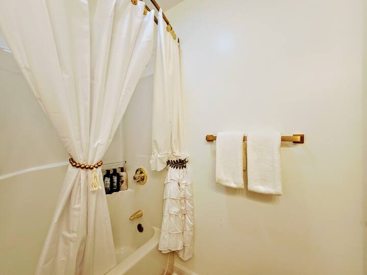 Bright, clean bathroom featuring a shower with elegant white curtains, neatly hung towels, and basic toiletries visible on the wall.