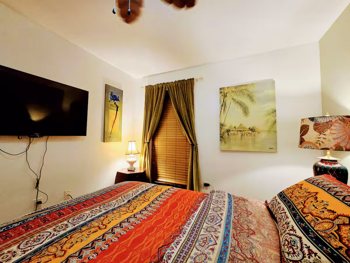 A warmly lit bedroom featuring a bed with colorful patterned quilts and pillows, positioned beneath a mounted television and framed tropical artwork on white walls. A small window with olive green curtains and wooden blinds allows soft light to filter through, complemented by a bedside lamp.