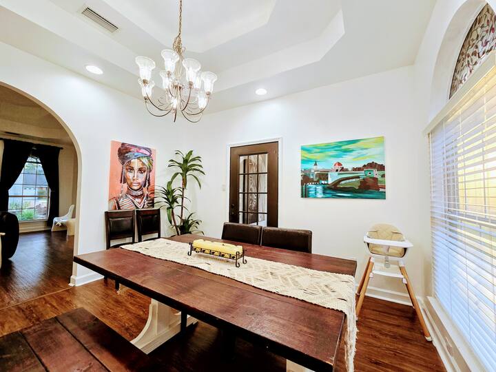 Bright dining area featuring a large wooden table, elegant chandelier, and colorful wall art, creating a warm, inviting atmosphere.