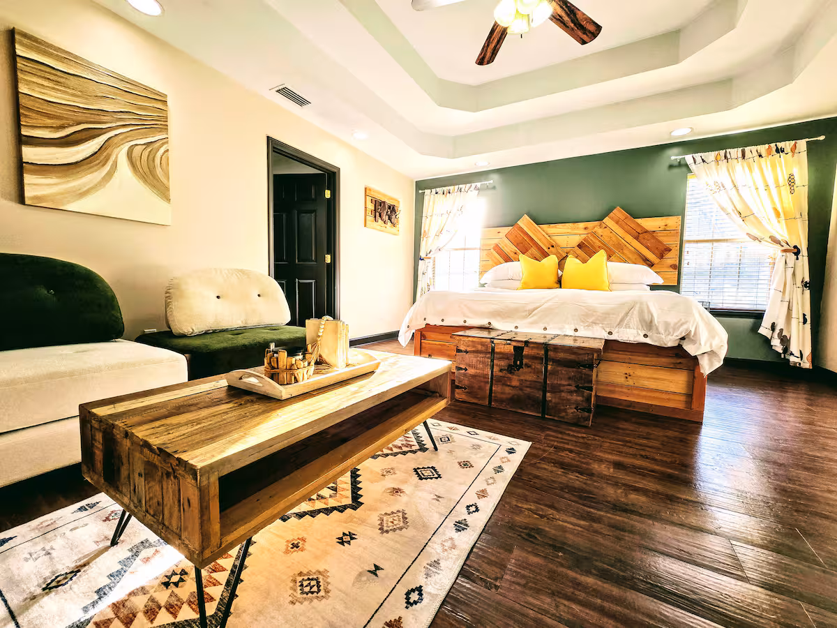 A spacious bedroom and living area features dark wood floors, a large wooden coffee table on a geometric rug, and a bed with a rustic wooden headboard against a green accent wall. A white and green sectional sofa sits on the left, and natural light streams through two windows dressed with patterned curtains.