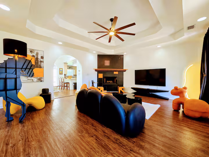 A wide-angle living room view featuring a multi-tiered tray ceiling with a large ceiling fan, a unique black horse-shaped floor lamp on the left, an arched entryway leading into the kitchen, a black-and-orange corner fireplace, a curved black sectional sofa in the foreground, a large flat-screen television on the right, and playful orange accent seating.
