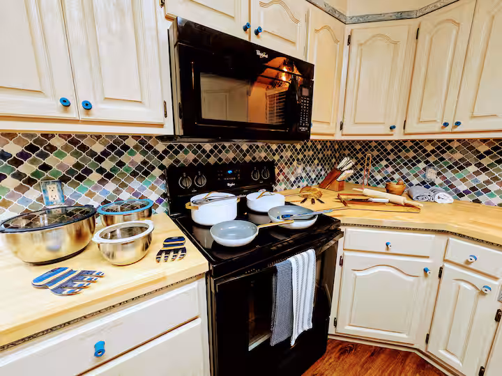 A kitchen cooking area featuring cream-colored raised-panel cabinets with blue knobs, a black microwave and electric range, a colorful lantern-pattern mosaic tile backsplash, and light wood countertops filled with cookware and mixing bowls.