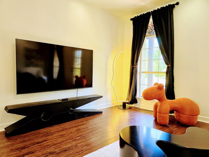Modern living room featuring a large flat-screen TV mounted on a black, angular console. A whimsical orange camel-shaped chair adds playful decor, illuminated by natural light from a nearby window.