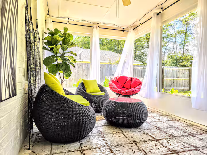 A bright sunroom features tiled flooring, large windows with sheer white curtains, and a mix of dark wicker chairs with bright lime green and red cushions, creating a cheerful and relaxing lounge space.