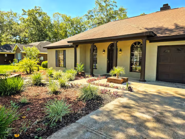 A single-story house with a tan exterior, dark roof, and dark brown garage door features arched windows and a covered front porch supported by dark pillars.