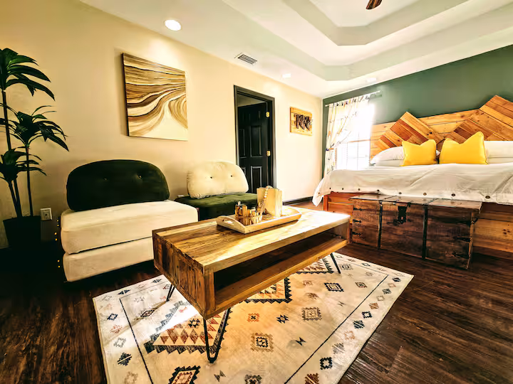 A spacious bedroom featuring a rustic wooden headboard and a platform bed with white linens and yellow pillows. In the foreground, a wooden coffee table sits on a patterned rug, facing a cream and dark green armchair and an adjacent matching stool.