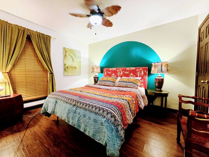 Colorful bedroom featuring a patterned bedspread, tropical decor, and a bold teal accent wall. Warm lighting creates a cozy atmosphere.