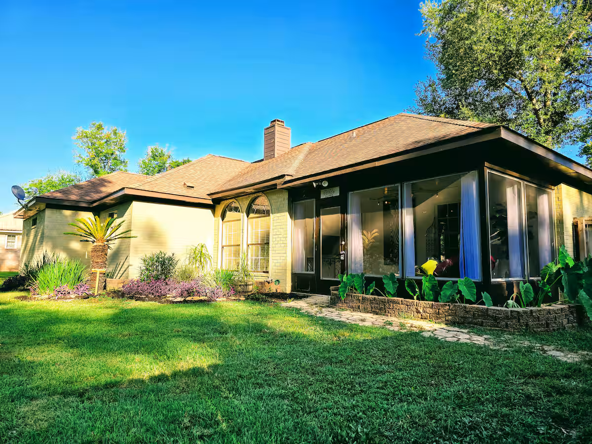 A single-story ranch-style home featuring light-colored brick walls, a dark-trimmed sunroom with large windows, and a brown shingled roof under a bright blue sky.