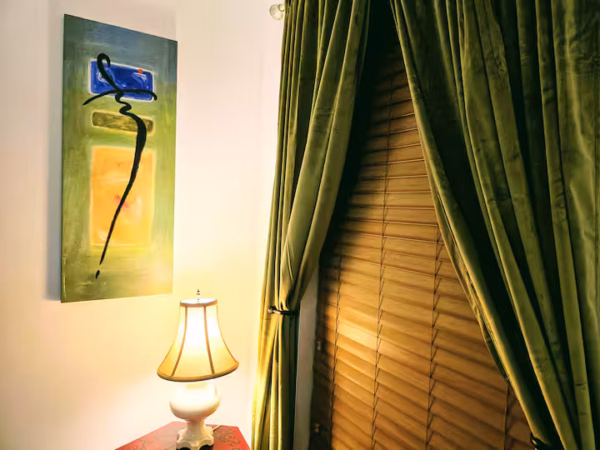 An interior shot features a lit white lamp on a table next to a wall with an abstract green painting. To the right, heavy olive green velvet curtains frame a window covered by wooden blinds.