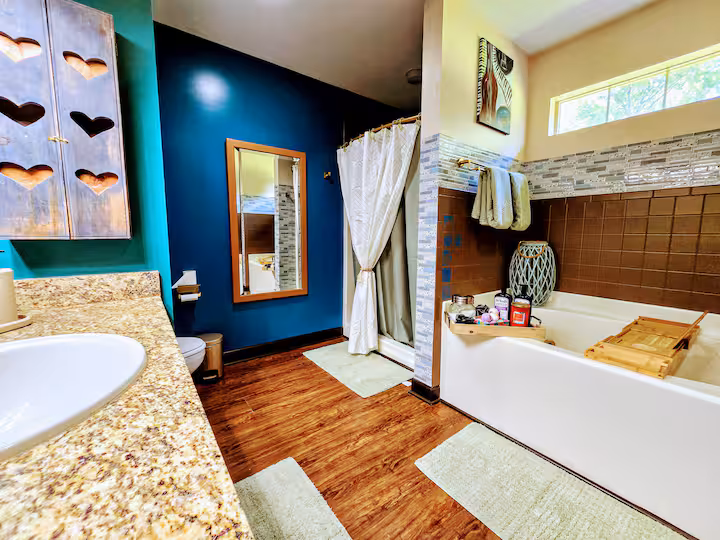 A spacious bathroom features dark blue and teal walls, rich brown tile, and a wooden floor.
