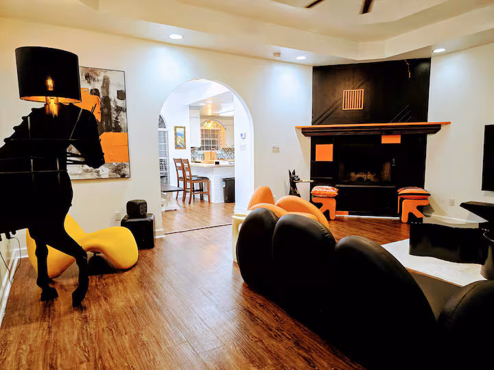 A wide-angle living room view featuring a unique black horse-shaped floor lamp on the left, a bold black and orange fireplace accent wall on the right, modern curved seating including a black sectional sofa and a bright yellow lounge chair, and an arched entryway leading into a bright kitchen.