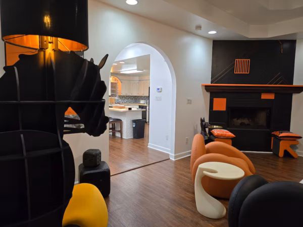 A living room view looking past a black horse-shaped floor lamp through an arched entryway into a kitchen, featuring a black-and-orange fireplace accent wall and modern curved seating.