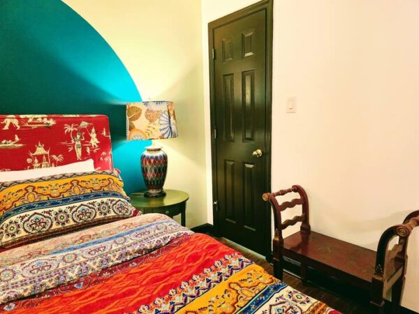 Colorful bedroom featuring a patterned bedspread, decorative lamp, and a dark door. A small wooden chair adds to the cozy ambiance.