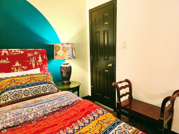 Colorful bedroom featuring a patterned bedspread, decorative lamp, and a dark door. A small wooden chair adds to the cozy ambiance.