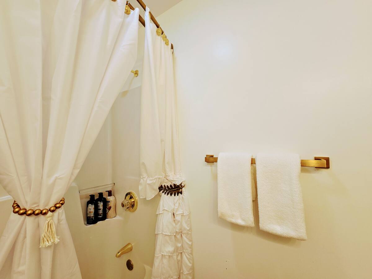 Bright, white bathroom featuring a decorative shower curtain, a gold towel rack with two white towels, and shampoo bottles displayed.