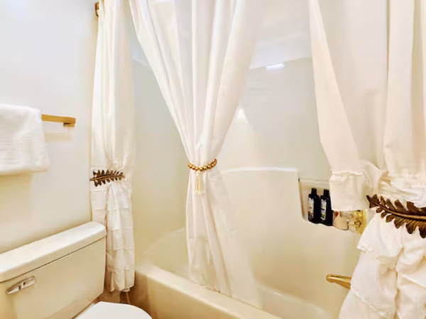 A well-lit bathroom featuring a white bathtub and shower surround, partially obscured by elegant white shower curtains gathered with decorative tiebacks.