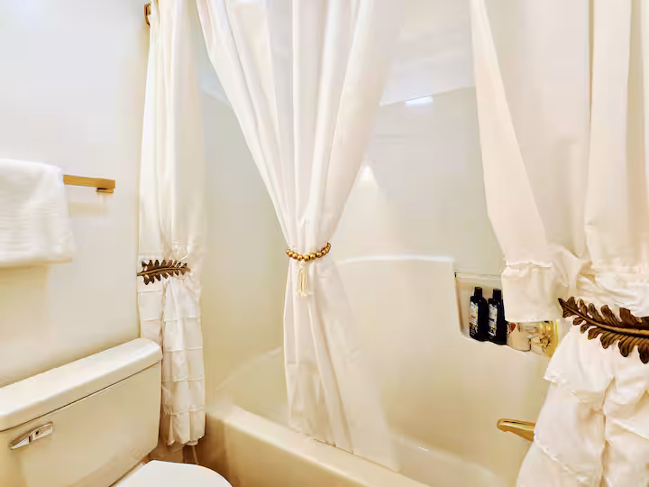 A well-lit bathroom featuring a white bathtub and shower surround, partially obscured by elegant white shower curtains gathered with decorative tiebacks.