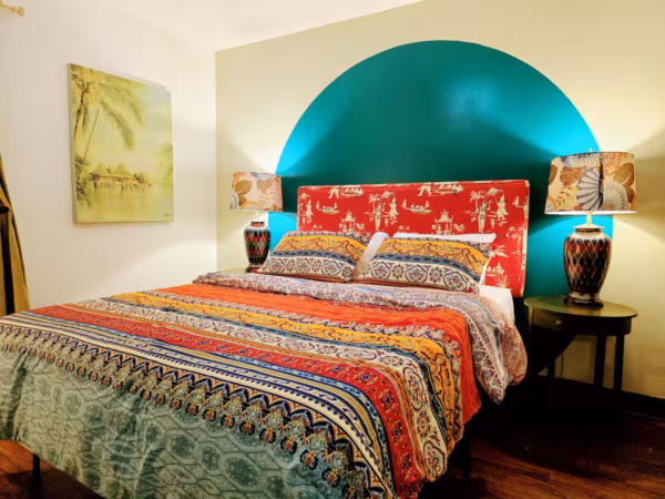 A cozy bedroom features a bed with a colorful patterned comforter and a red, patterned headboard against a wall with a teal semi-circle painted behind it. Matching lamps with patterned shades sit on small dark wood nightstands on either side of the bed, and a painting of a tropical stilt village hangs on the white wall to the left.
