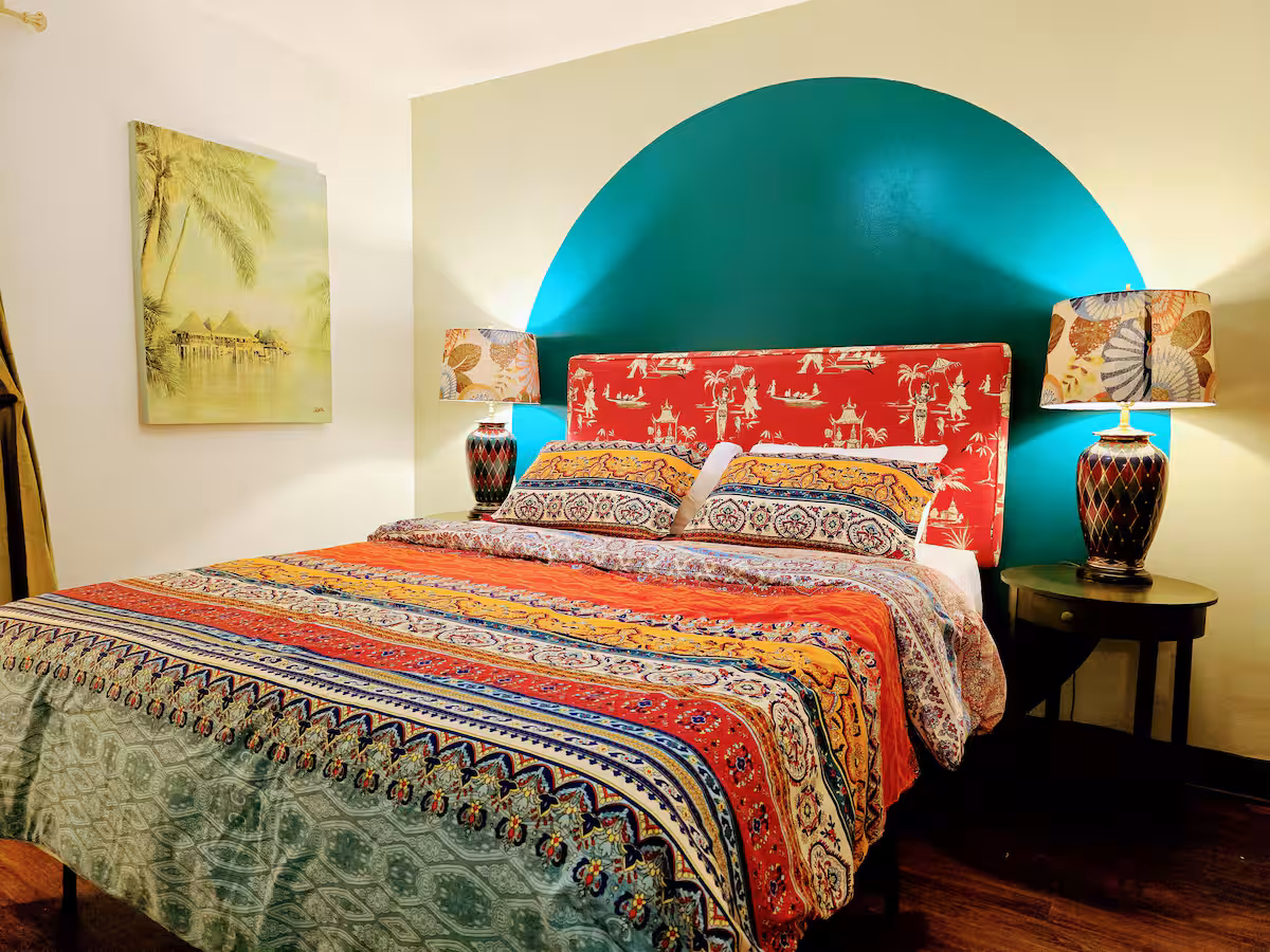 A cozy bedroom features a bed with a colorful patterned comforter and a red, patterned headboard against a wall with a teal semi-circle painted behind it. Matching lamps with patterned shades sit on small dark wood nightstands on either side of the bed, and a painting of a tropical stilt village hangs on the white wall to the left.