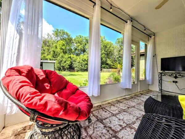 Bright sunroom featuring a vibrant red cushioned chair, floor-to-ceiling windows with white drapes, and a view of a green backyard.