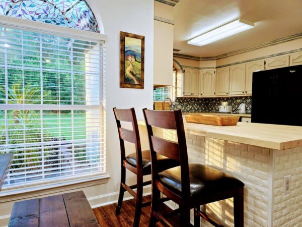 Bright kitchen with a wooden breakfast bar, two dark chairs, and a stained glass window. A serene garden view is visible through the blinds.