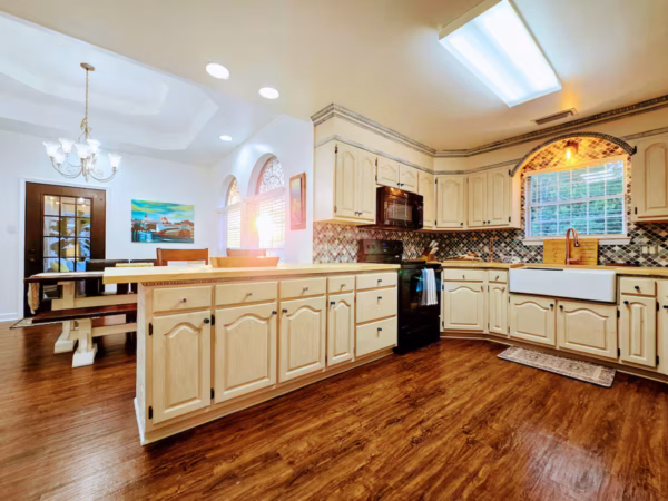 A spacious traditional kitchen featuring cream-colored raised-panel cabinetry, a large kitchen island with light wood countertops, rich hardwood flooring, a farmhouse sink beneath a decorative arched window with a patterned tile backsplash, and an adjacent dining area.