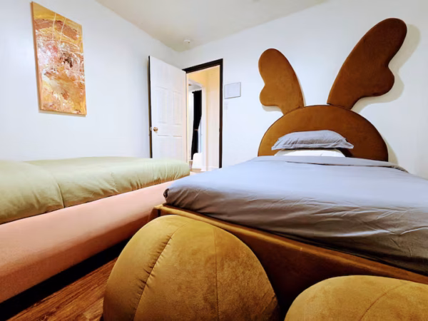 A cozy bedroom features a unique bed with a brown upholstered headboard shaped like rabbit ears. A second bed with a green comforter is on the left, and a gold beanbag sits in the foreground on a wooden floor.