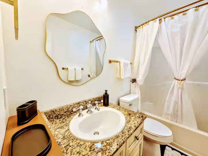 An interior shot of a brightly lit bathroom featuring a speckled granite vanity with a white undermount sink, a brass-framed irregular-shaped mirror, and a white toilet next to a bathtub with white curtains.